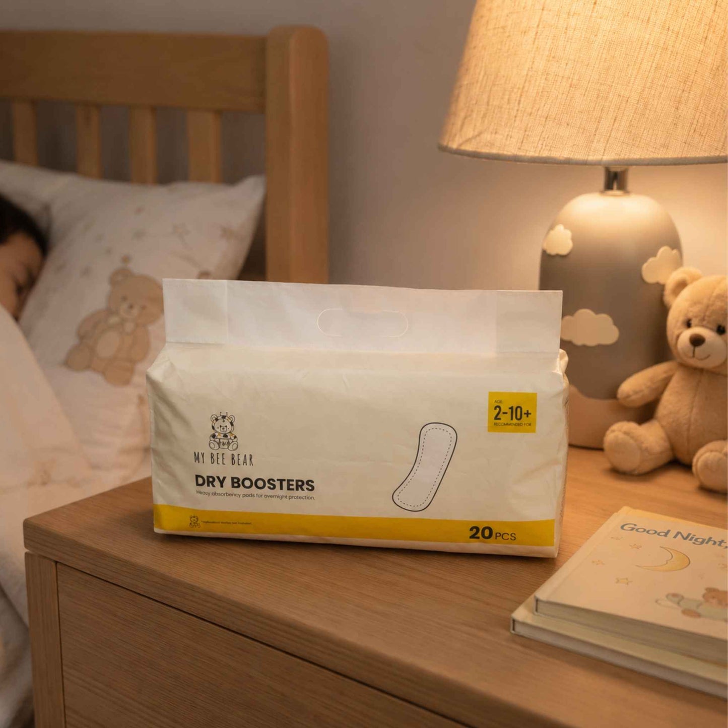 A package of My Bet Bear Dry Boosters hand warmers sits on a wooden bedside table next to a bed, with a lit lamp and a teddy bear in the background.
