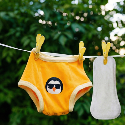 Yellow children's underwear with a penguin design hanging on a clothesline against a green leafy background.