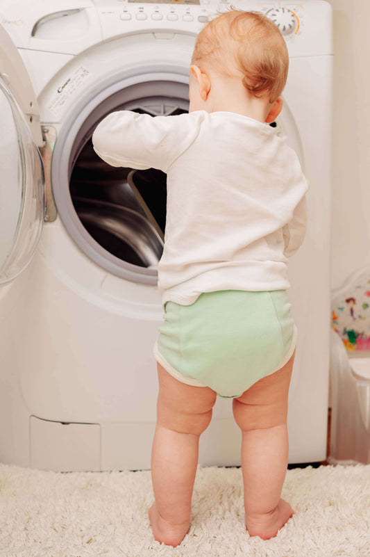 Baby in a potty training underwear standing in front of an open washing machine.