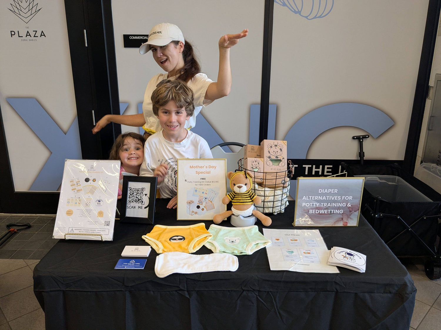 Woman and children with a table displaying products and certificates in front of a building entrance.