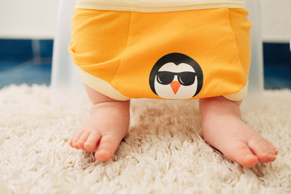 Baby wearing a yellow diaper with a penguin design on a white carpet.