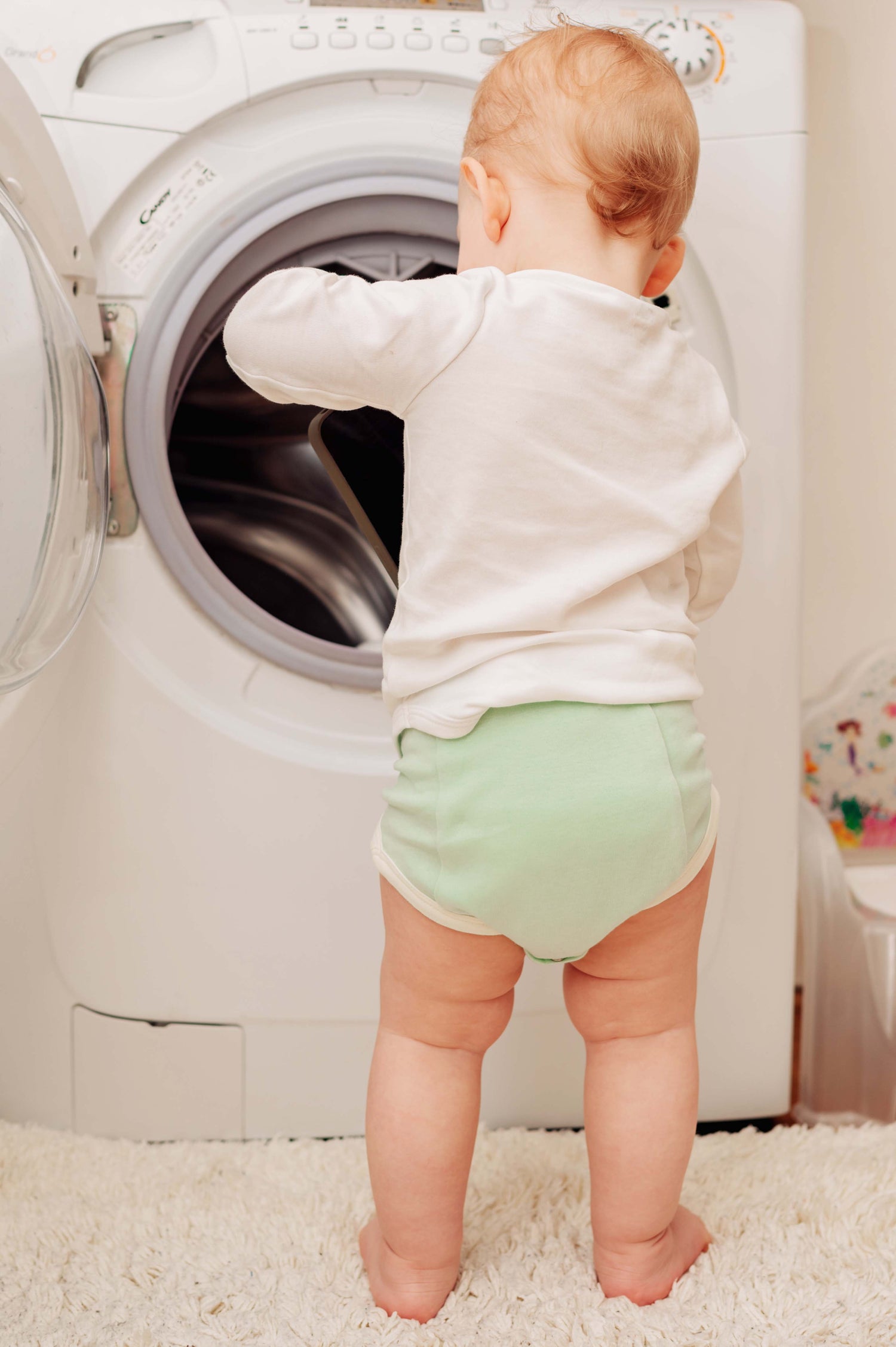 Baby in a diaper standing in front of an open washing machine.